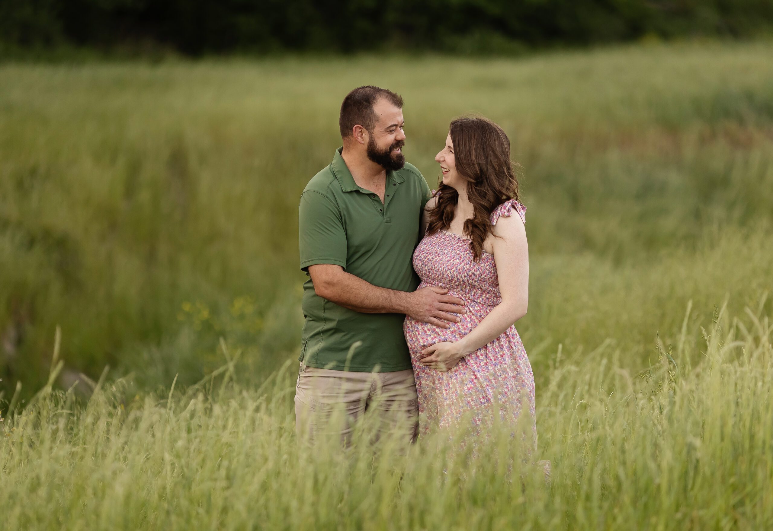 Expectant couple standing in a lush green field during an outdoor maternity photo session, with the man in a green polo shirt gently holding the woman’s pregnant belly as they smile at each other.