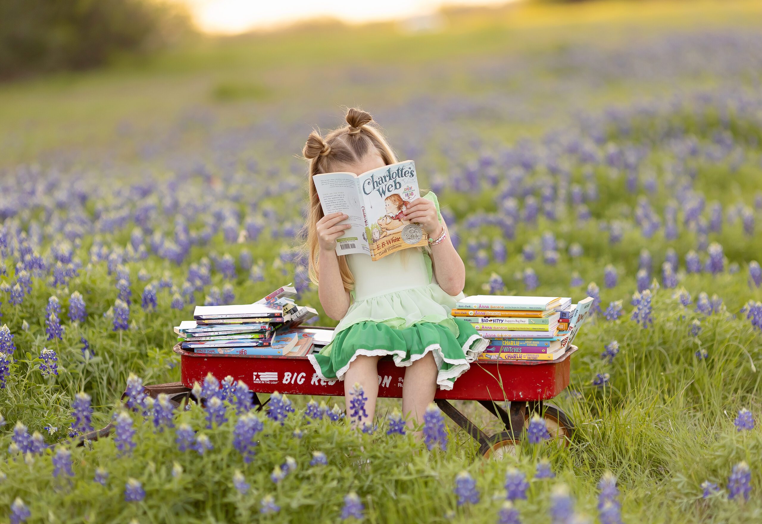 Young girl in a green ruffle dress reading “Charlotte’s Web” while sitting on a red wagon surrounded by Texas bluebonnets and stacks of books.