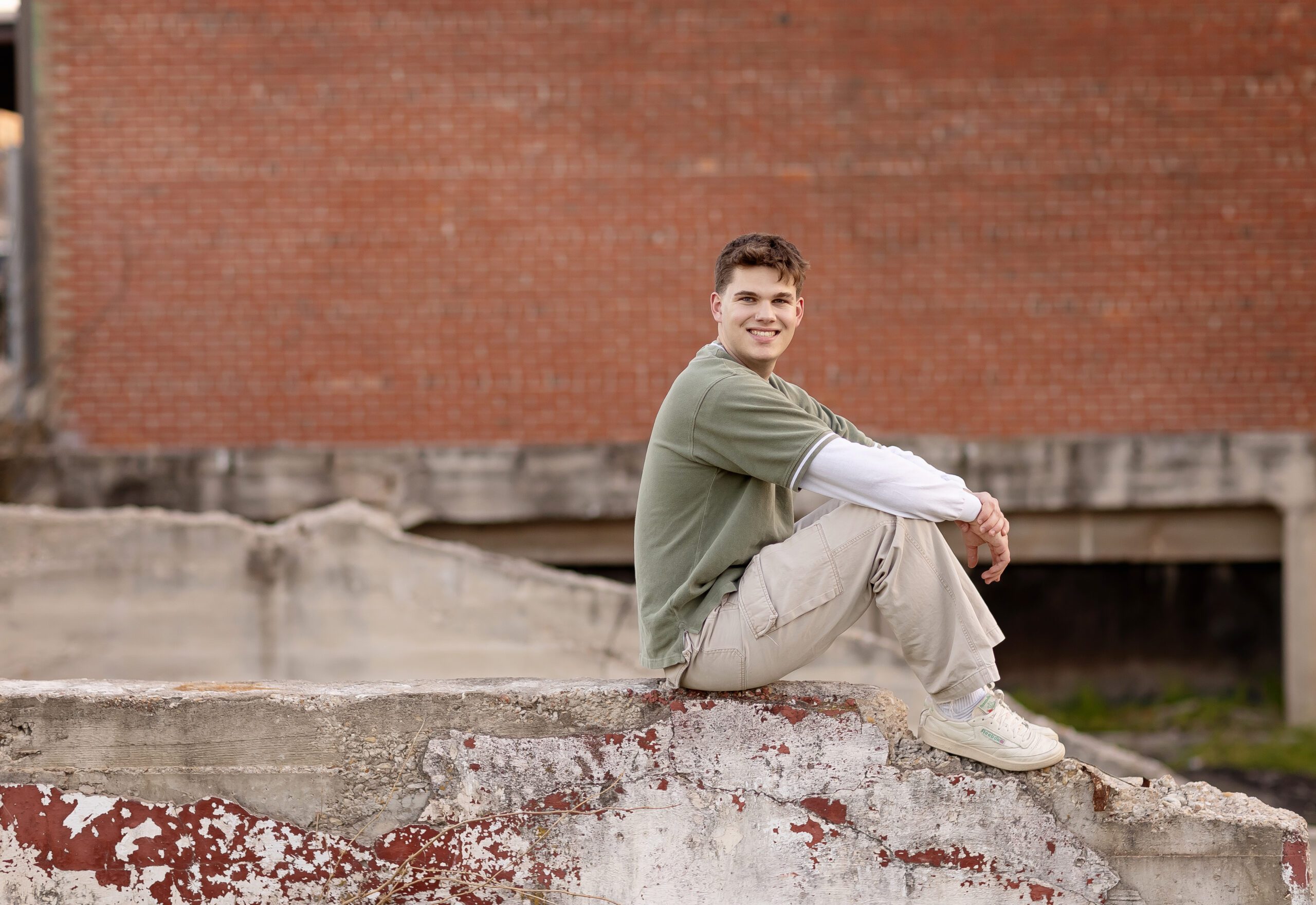 High school senior boy sitting on a concrete wall in front of a red brick building, wearing a green shirt and khaki pants, smiling at the camera.