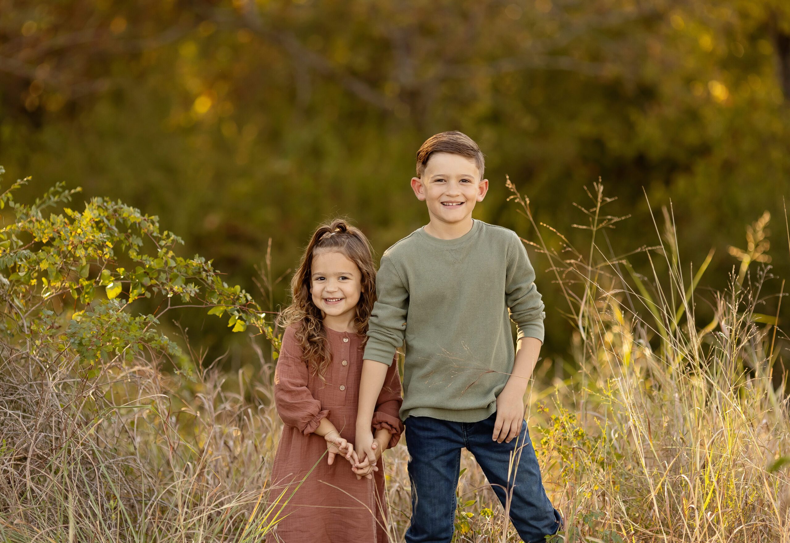 Brother and sister holding hands and smiling during the Dewsnap family photo session in a tall grass field near the George Bush Library in College Station, TX.