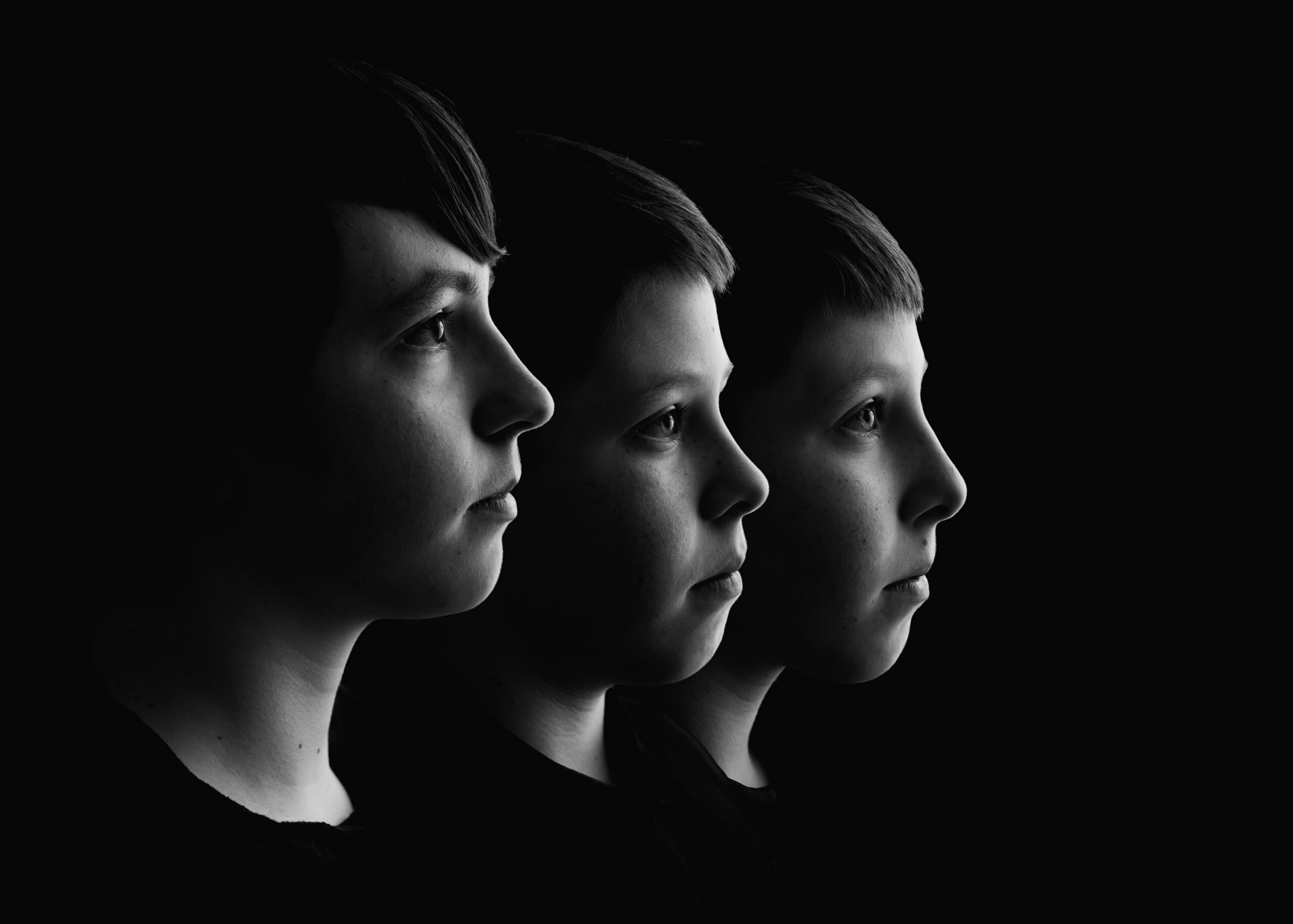 Three siblings photographed in side profile with soft sculpted lighting on a black background.